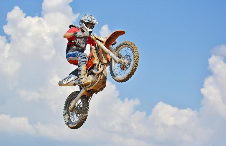 Motocross rider performs an airborne jump under a bright blue sky with clouds.